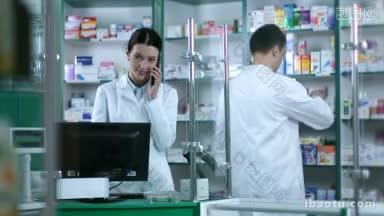 漂亮的女药剂师在桌子后面用电脑显示器<strong>接电话</strong>，男同事检查货架上的药品